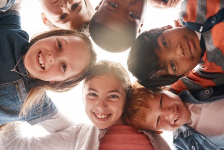 6 children from different races stood in a circle and looking down into the camera positioned on the ground facing up.