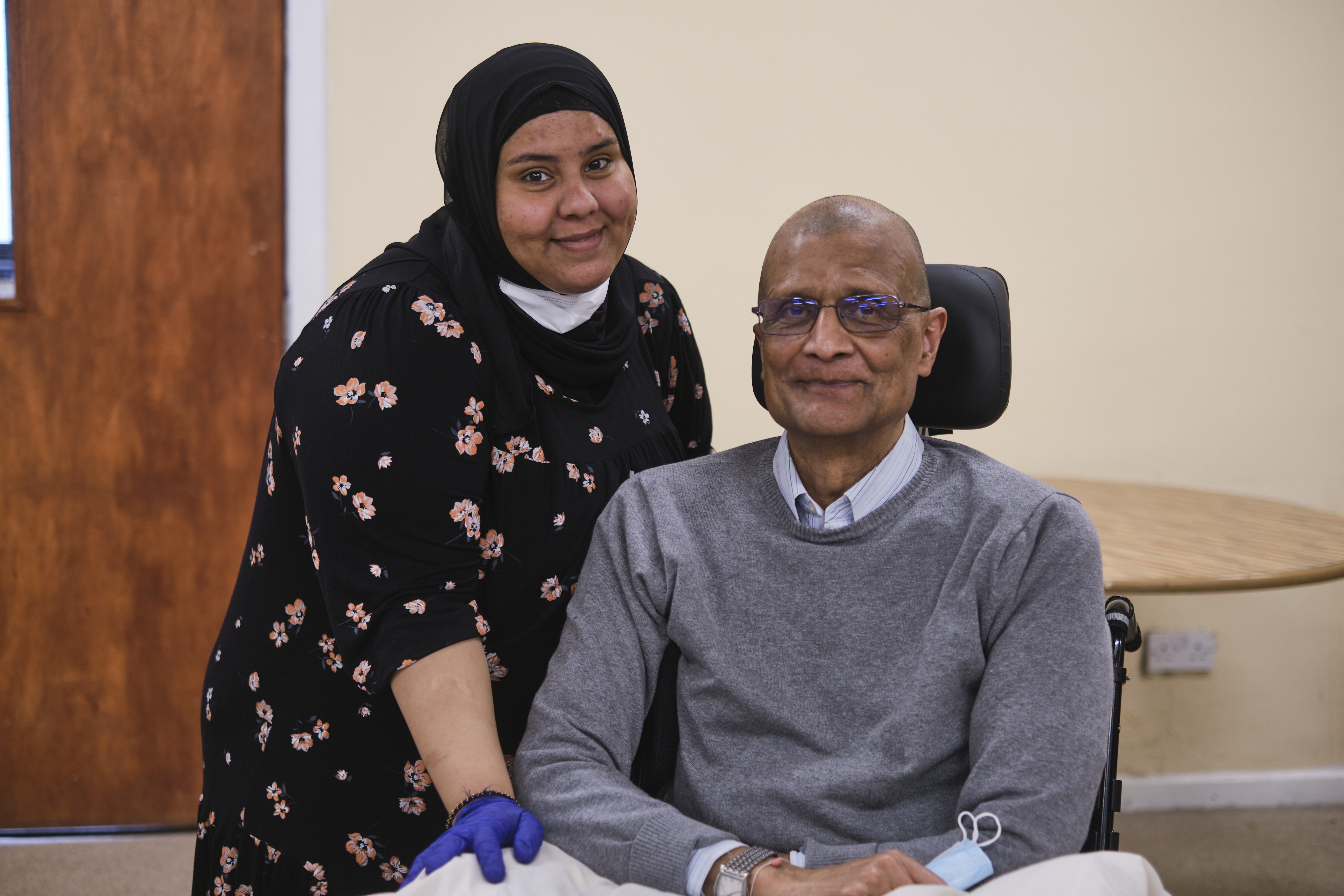 A woman wearing a headscarf, black flowery dress and blue medical gloves is stood next to an older man in a wheelchair wearing a grey jumper and holding a face mask. Both are smiling at the camera.